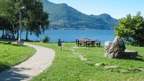 A park with a paved path, benches, and a picnic table by a lake with mountains in the background. - Apartment by Lake Como with Scenic Views (Dervio)