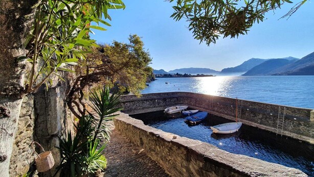 A stone wall with boats docked in a calm body of water, surrounded by greenery and mountains in the background. - Apartment by Lake Como with Scenic Views (Dervio)