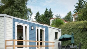 Une maison mobile dotée d’une terrasse, de meubles de jardin et offrant une vue sur les arbres et les maisons environnantes.