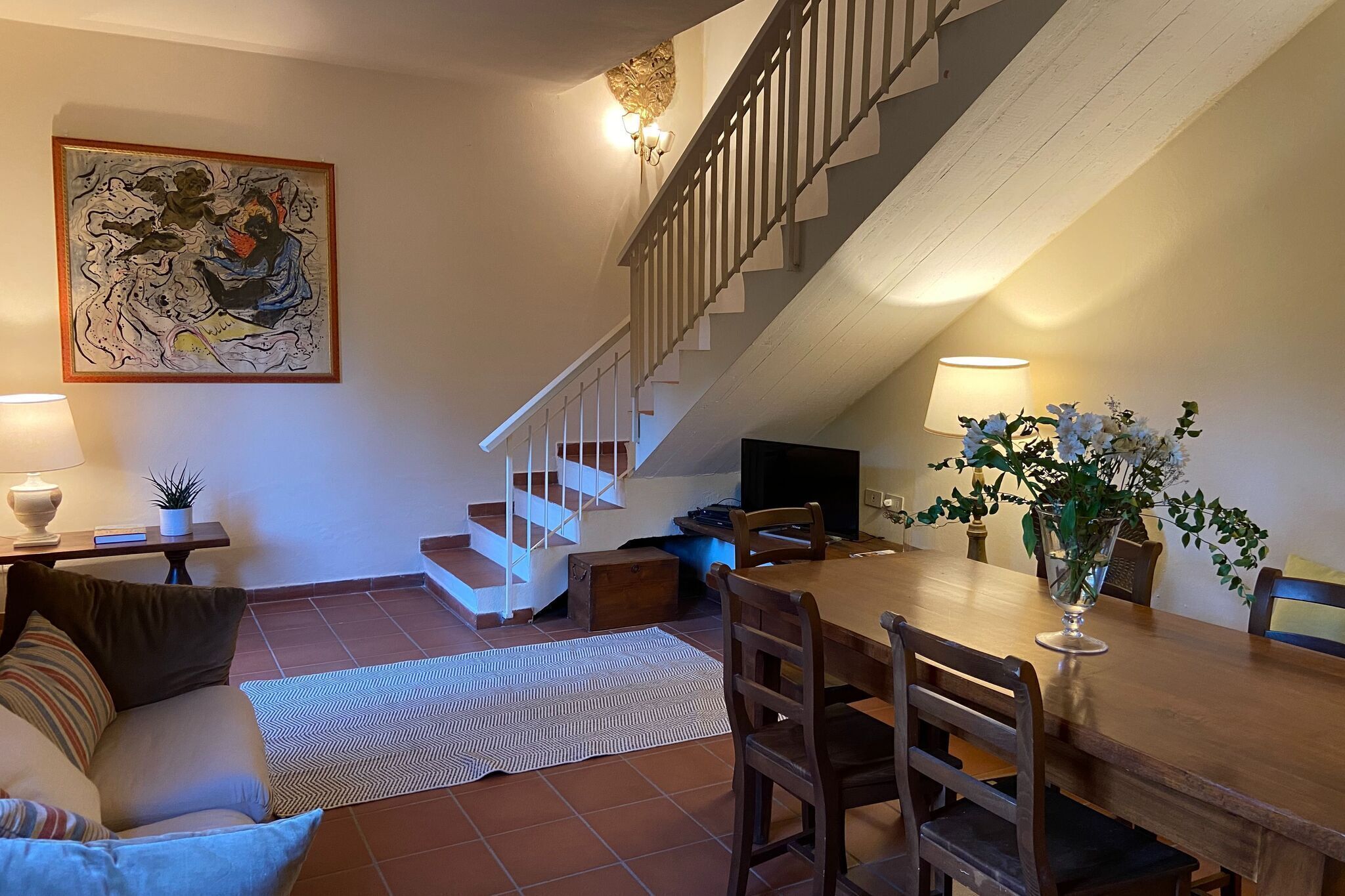 A dining area with a wooden table and chairs, a staircase, and a painting on the wall.