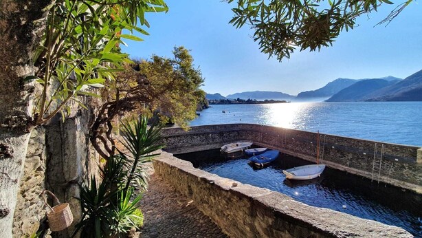 Un muro de piedra con barcos amarrados en aguas tranquilas, rodeado de vegetación y montañas al fondo.