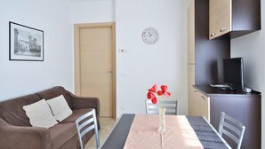 Un salón moderno con un sofá marrón, una mesa de comedor con un jarrón de flores rojas, un reloj en la pared y un televisor sobre un mueble.