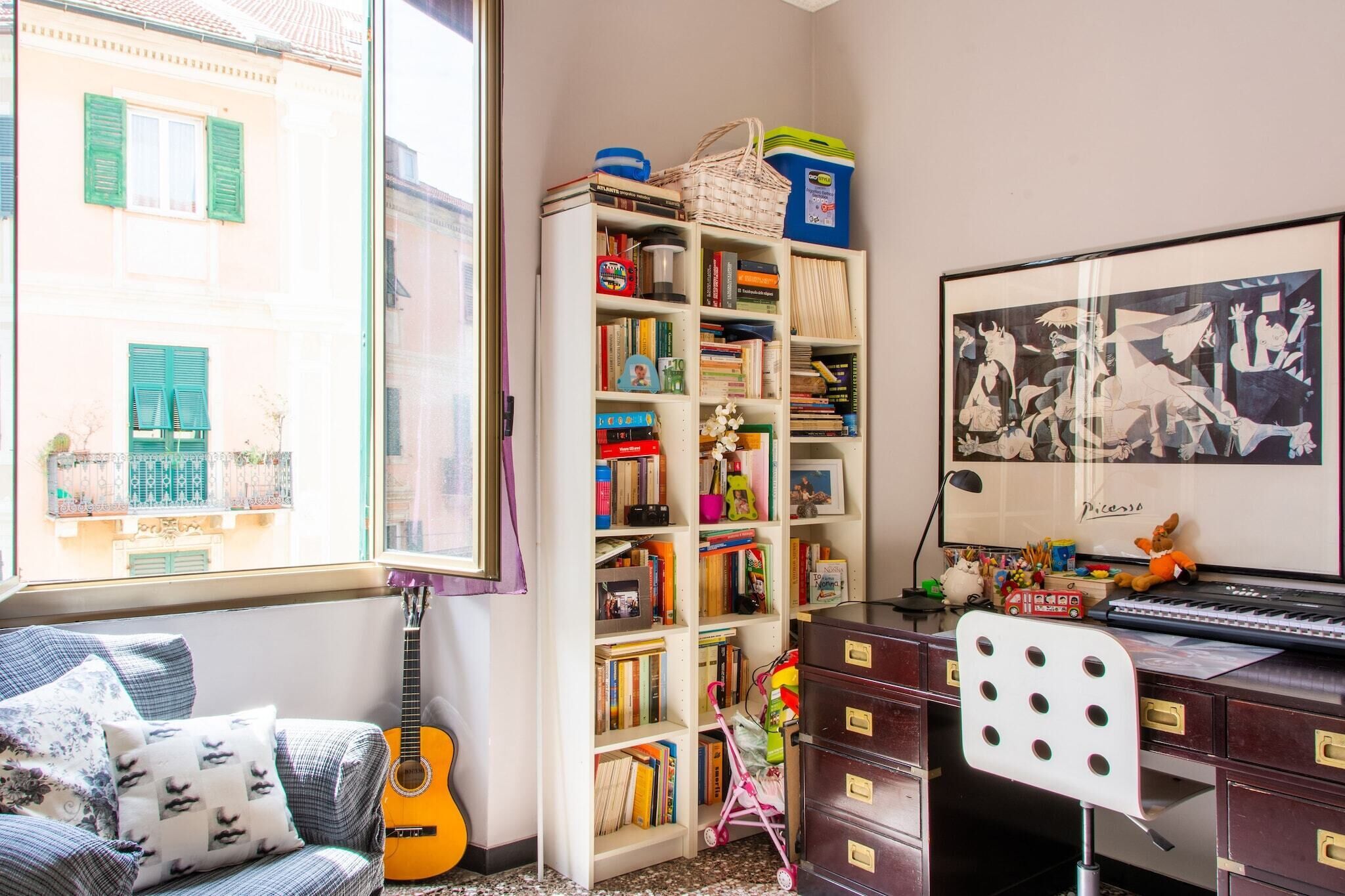 Ein gemütliches Zimmer mit Bücherregal, einer Gitarre, einem Schreibtisch mit Tastatur und einem Fenster mit Blick auf Gebäude.