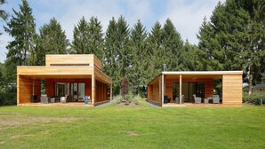 Une maison moderne à deux étages, dotée d’une façade en bois, de grandes fenêtres en verre et d’un espace extérieur aménagé avec un coin salon couvert.