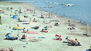 En strand med flera parasoller och solstolar, båtar i vattnet och fri utsikt över havet.