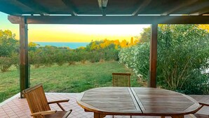 Une terrasse couverte, avec une table et des chaises en bois, donnant sur un espace verdoyant agrémenté d’arbres et offrant une vue sur la mer.