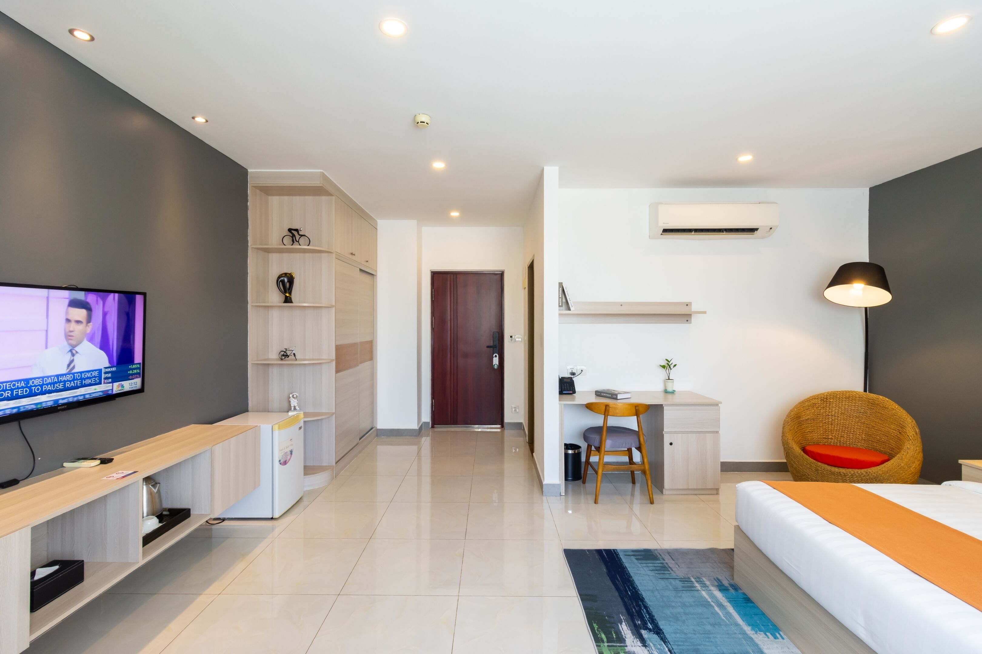 A modern hotel room with a TV, a desk, a chair, a bed, and a small kitchenette.