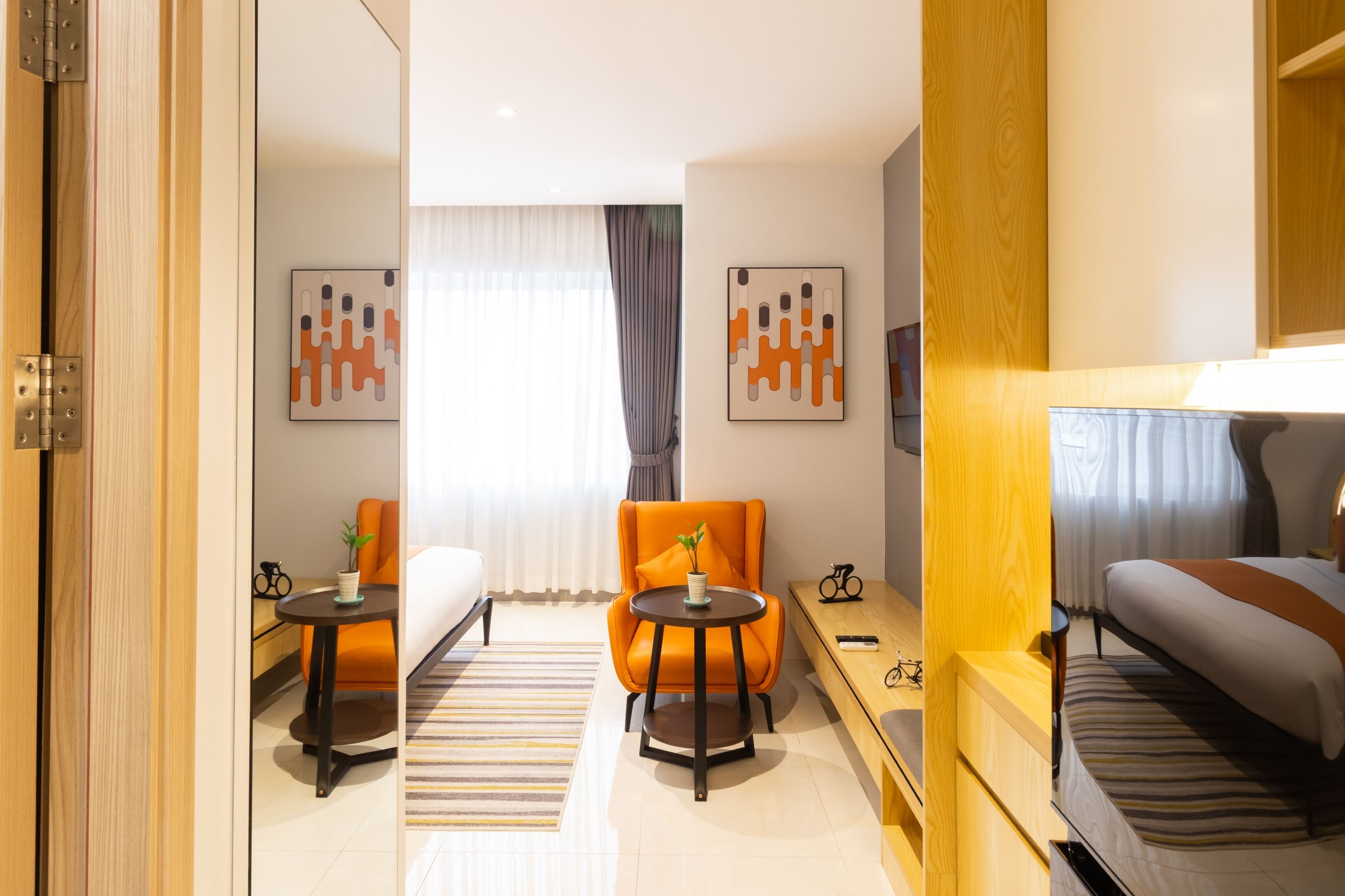 A modern hotel room with a bed, a small table with a plant, an orange chair, and a sofa. There are abstract art pieces on the walls and a window with curtains.