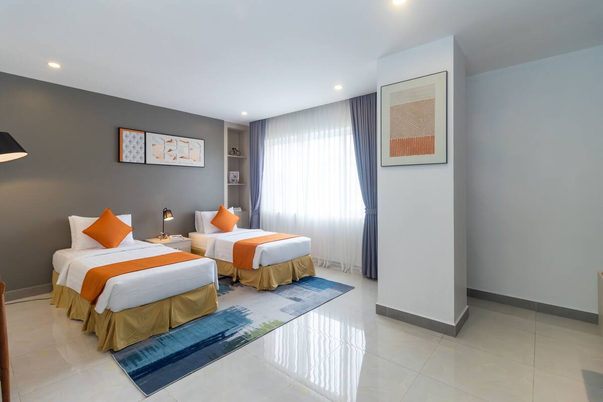 A hotel room with two beds, a large window with curtains, a framed artwork on the wall, and a blue rug.