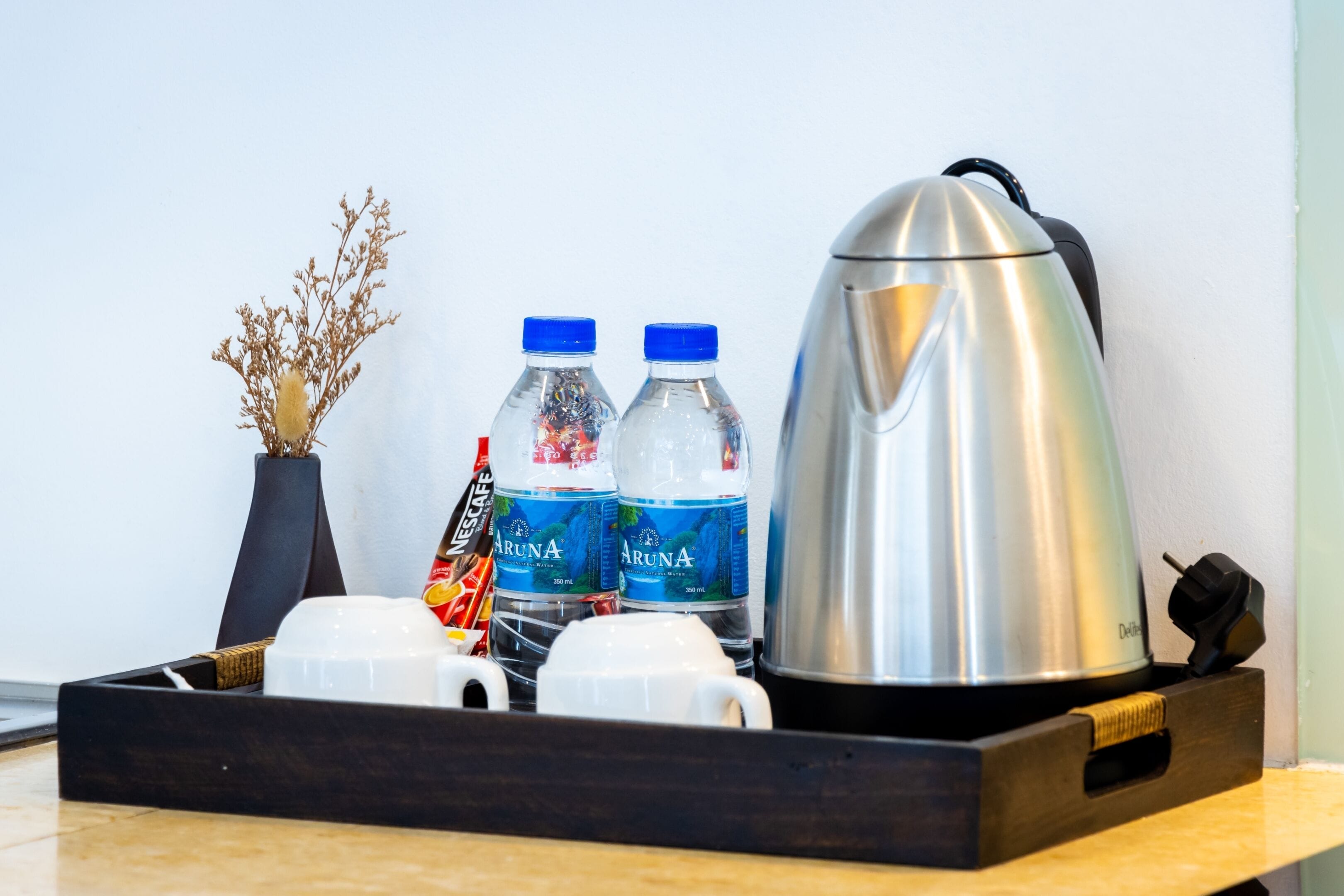 A tray with two bottles of water, two white cups, a black vase with dried flowers, and a silver electric kettle.