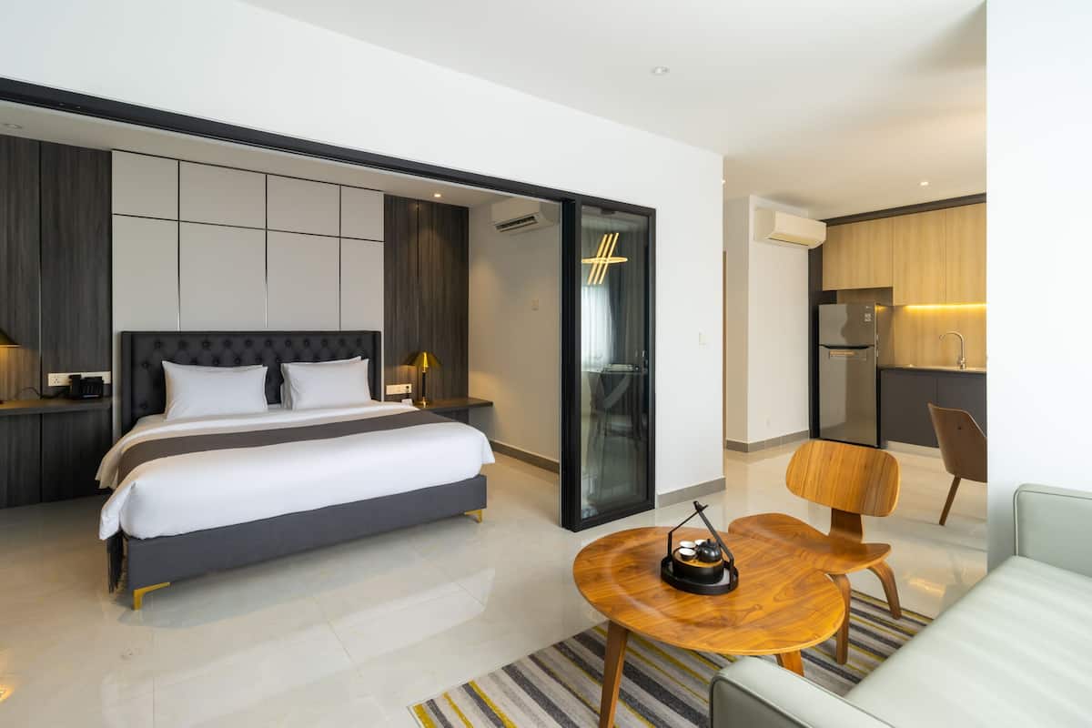 A modern hotel room with a large bed, a small dining table, a sofa, and a kitchen area.