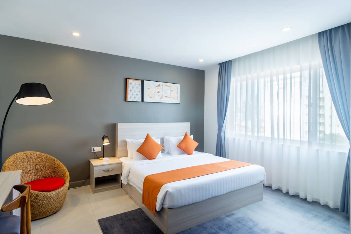 A modern hotel room with a large bed, a bedside table, a lamp, a wicker chair, and a window with sheer curtains.
