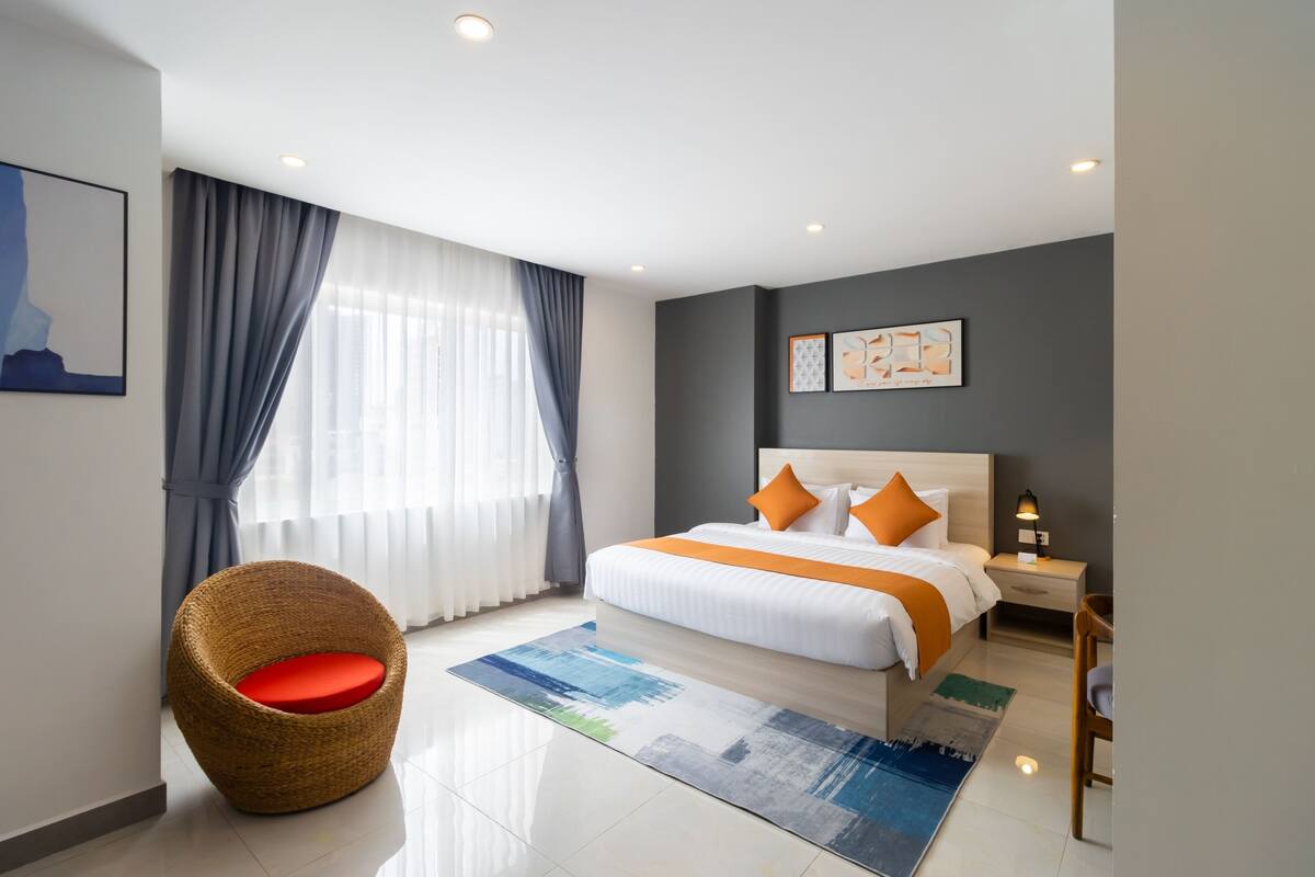 A modern hotel room with a large bed, a wicker chair, a nightstand, and a view of the city through the window.