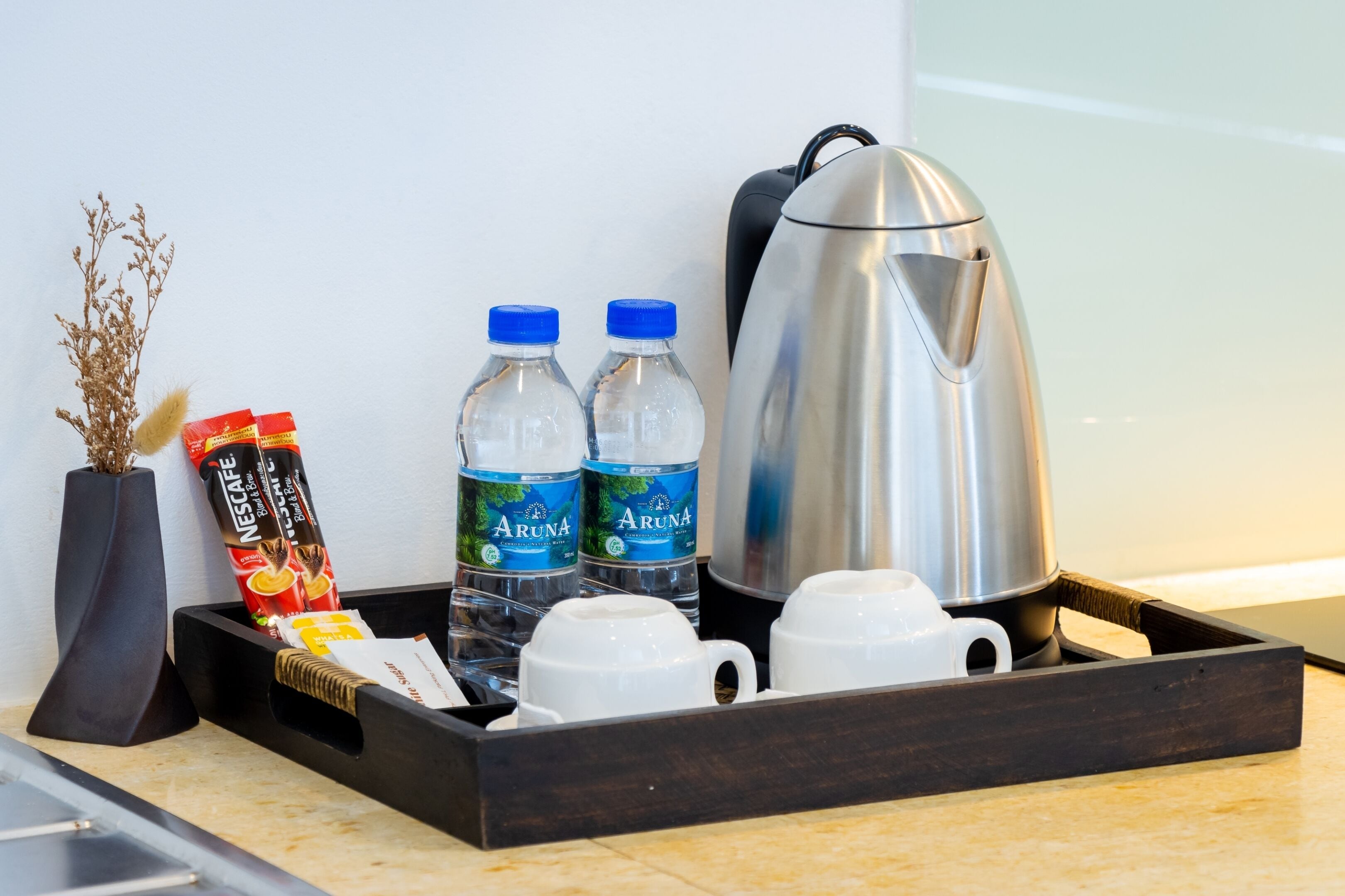 Una bandeja con un paquete de café Nescafé, dos botellas de agua Aruna, una tetera eléctrica plateada y tazas blancas sobre una encimera de cocina.