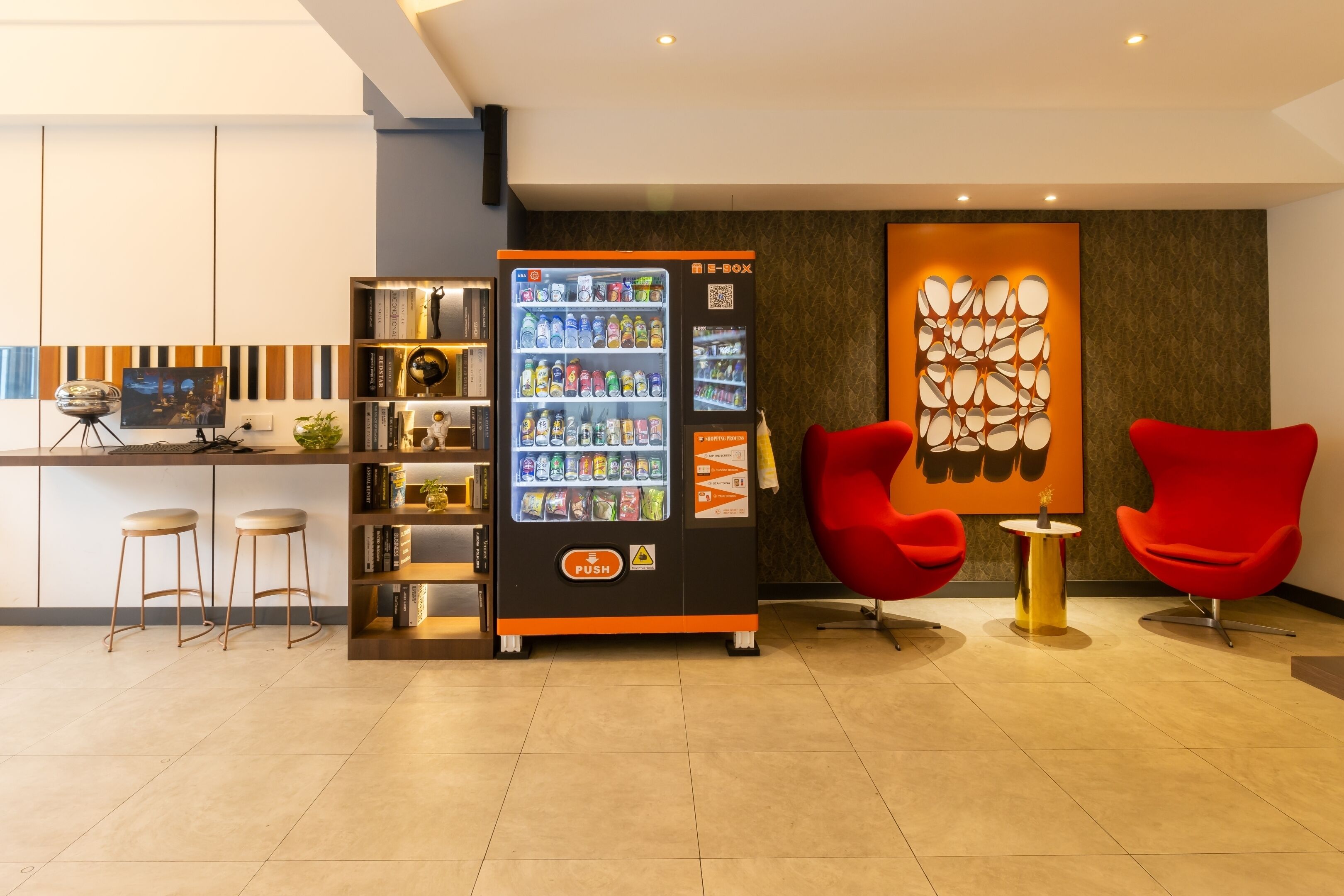 A modern hotel lobby with a vending machine, red chairs, a bookshelf, and a decorative wall.