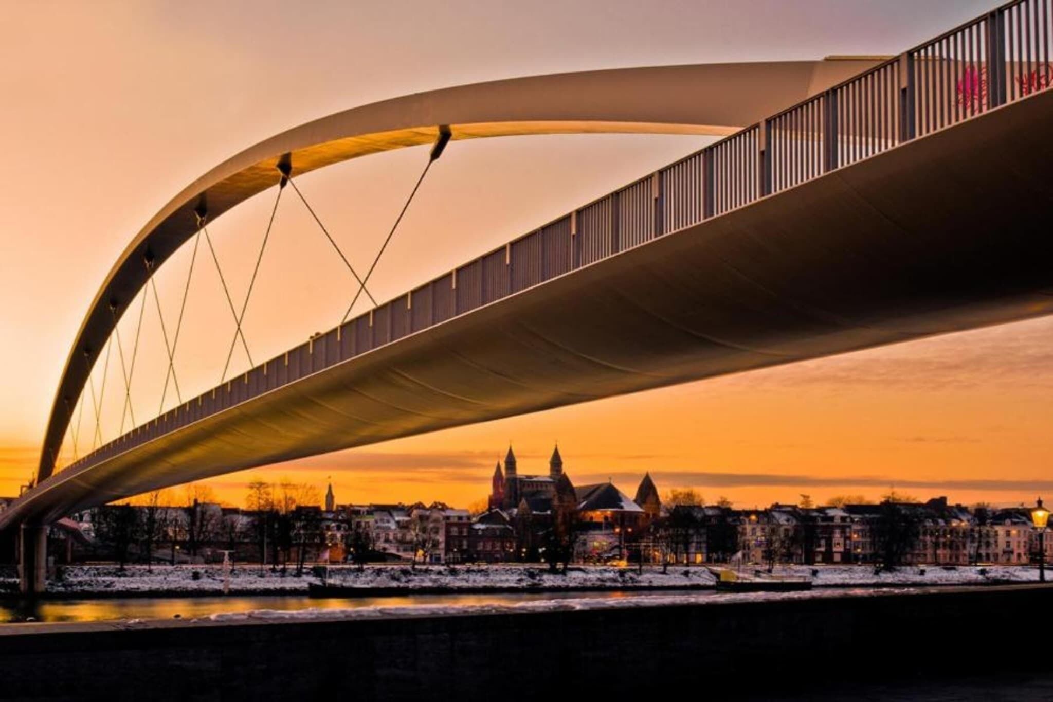 Un pont moderne au design incurvé enjambant une rivière, avec des bâtiments et une ville en arrière-plan.