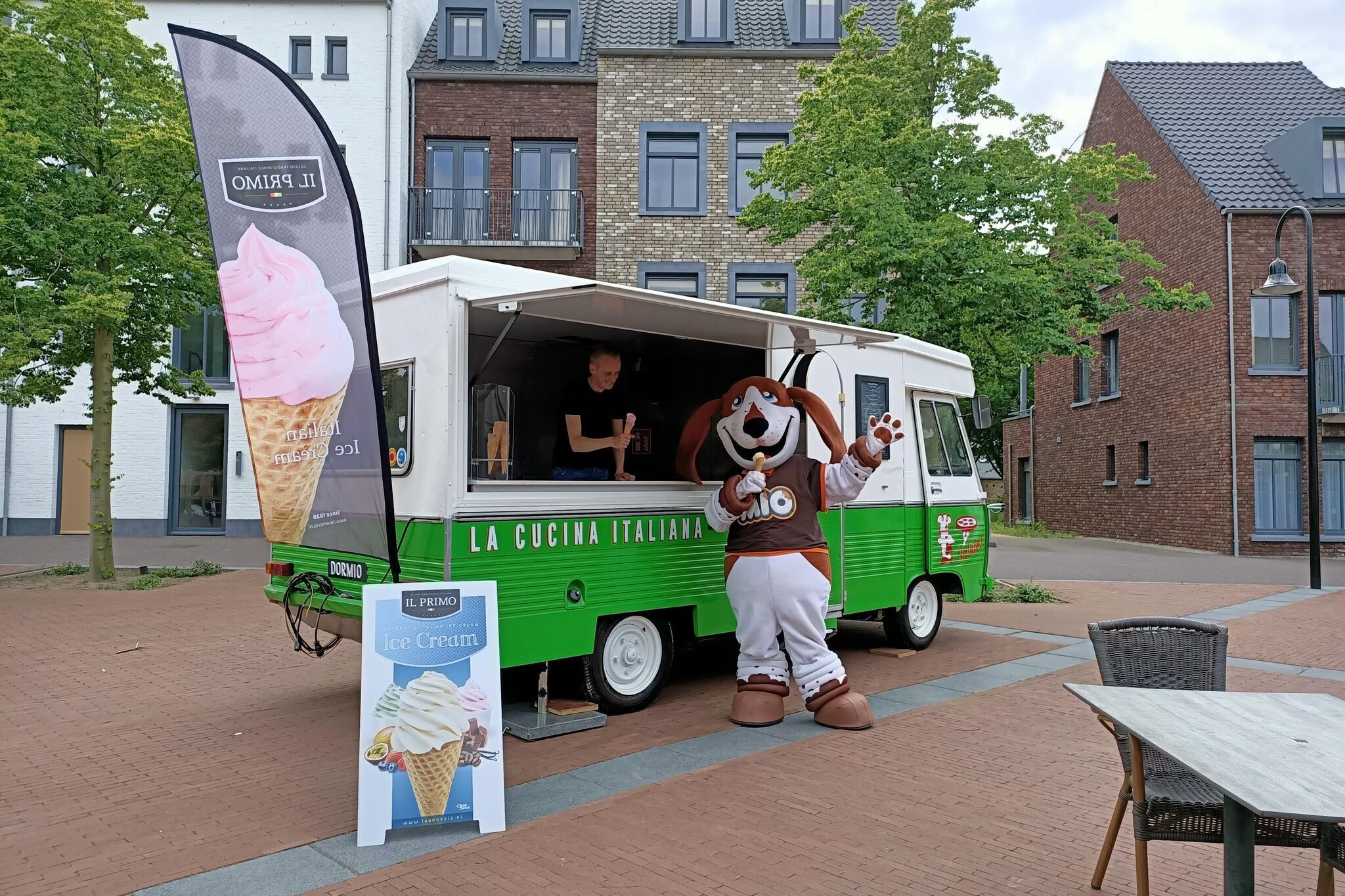 Un camion de glaces, orné d’une mascotte, est garé dans un espace extérieur, avec des bâtiments en briques en arrière-plan.