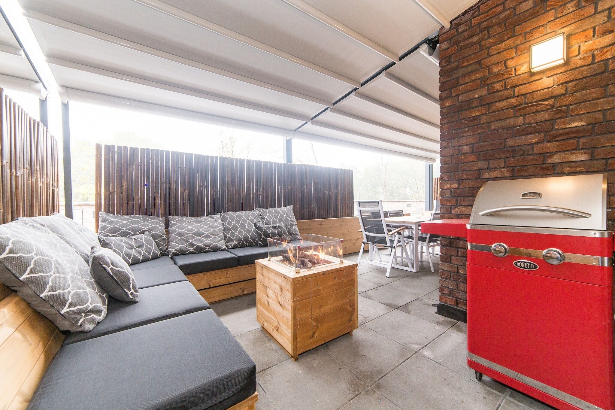 A covered outdoor area with a wooden sofa, a small fire table, and a red barbecue grill.