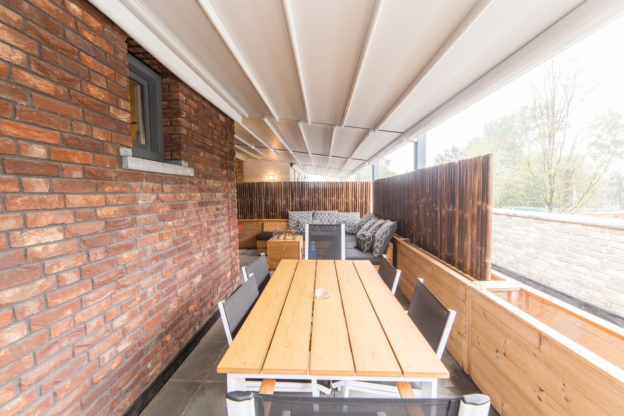 Un espacio al aire libre con mesas y bancos de madera, una pared de ladrillo y vistas del exterior.