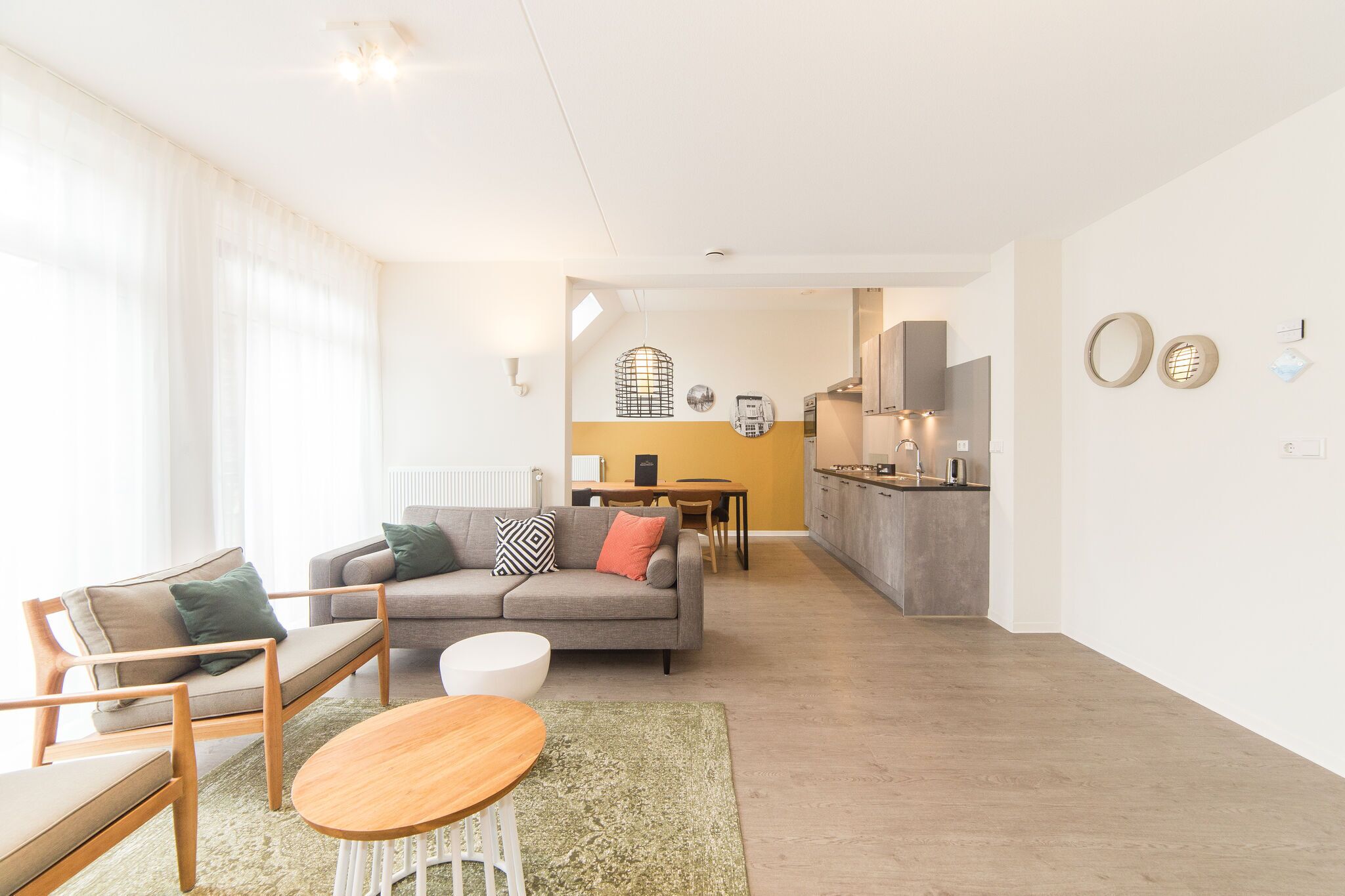 Een moderne woonkamer met een bank, stoelen en een eettafel. Achtergrond toont een keuken met een eettafel en stoelen.
