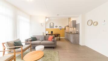 Een moderne woonkamer met een bank, stoelen en een eettafel. Achtergrond toont een keuken met een eettafel en stoelen.