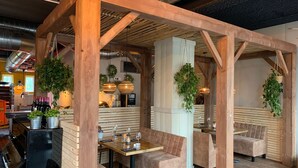 Un restaurant avec des poutres en bois, des sièges confortables, des plantes suspendues et une cuisine visible au fond de la salle.