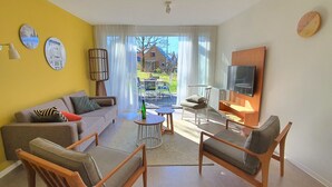 Een moderne woonkamer met een bank, stoelen, een televisie en uitzicht op een huis door grote ramen.