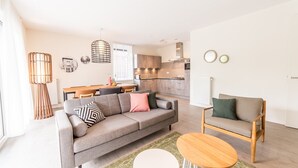 Een moderne woonkamer met een grijze bank, een houten salontafel en een houten fauteuil. Achtergrond toont een keuken met houten kastjes en een eethoek.