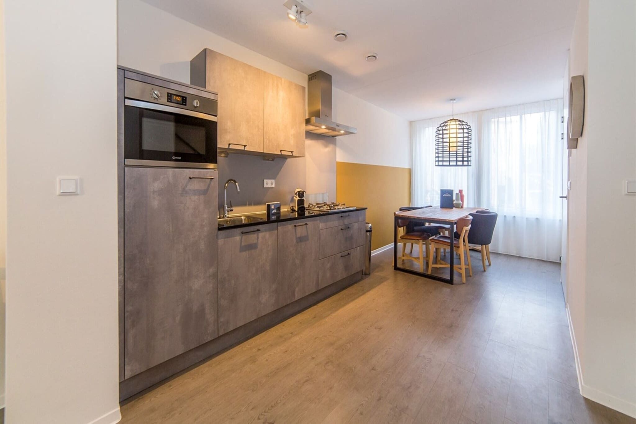 A modern kitchen with a built-in oven, microwave, and stainless steel range hood. There is a dining area with a table and chairs.