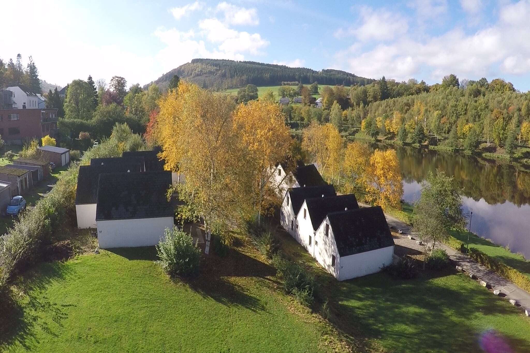 A row of white buildings with black roofs, a grassy area, and a lake with trees in autumn colors.