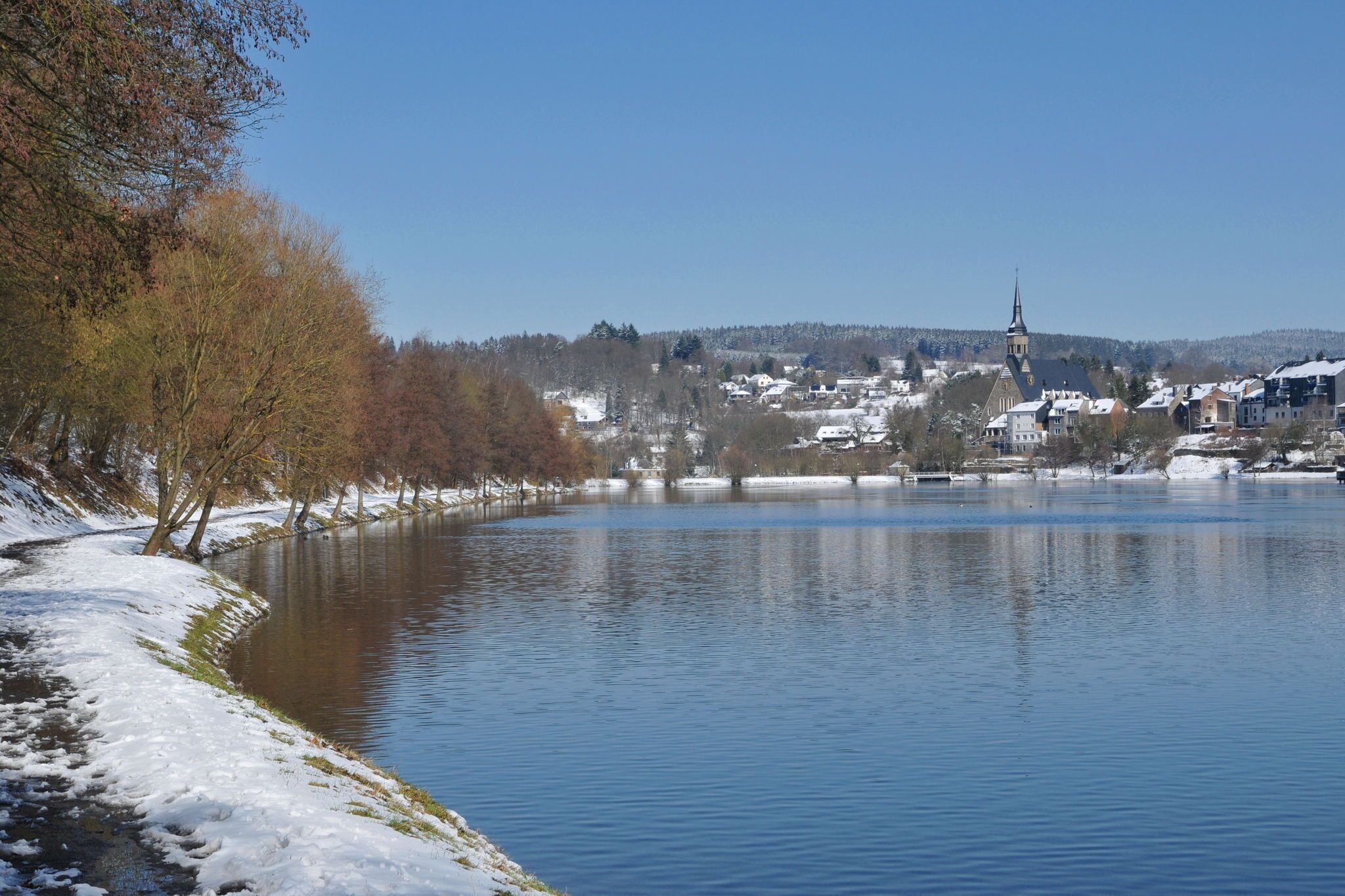 Une vue paisible sur un lac, avec le clocher d’une église qui se dessine à l’horizon, entouré d’arbres et de bâtiments recouverts de neige.