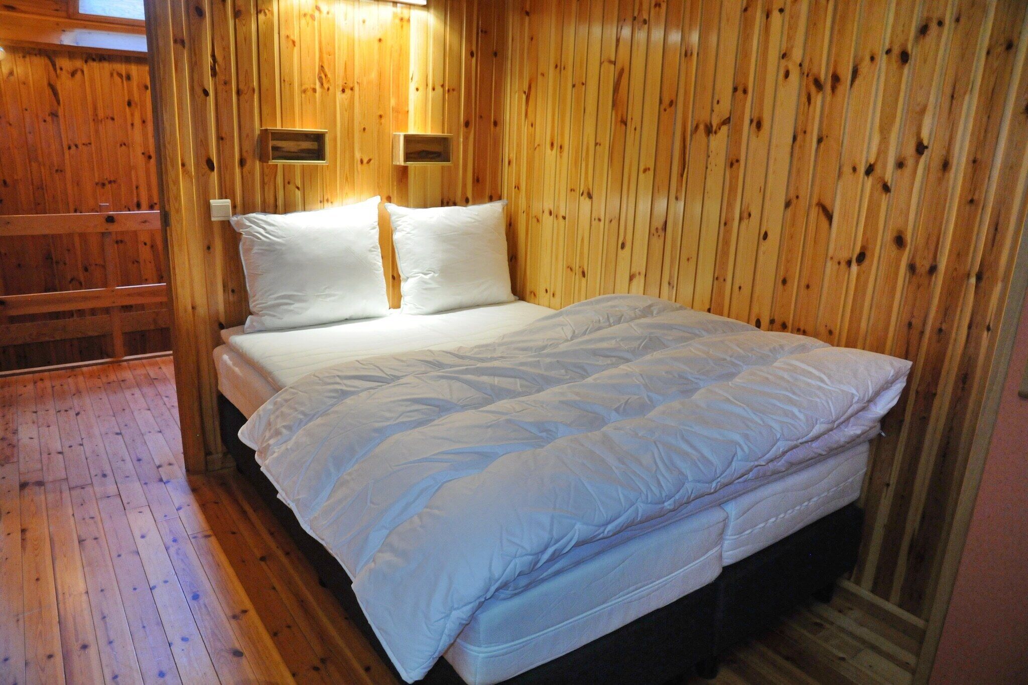 A wooden-paneled room with a bed, two wall-mounted light fixtures, and a wooden floor.