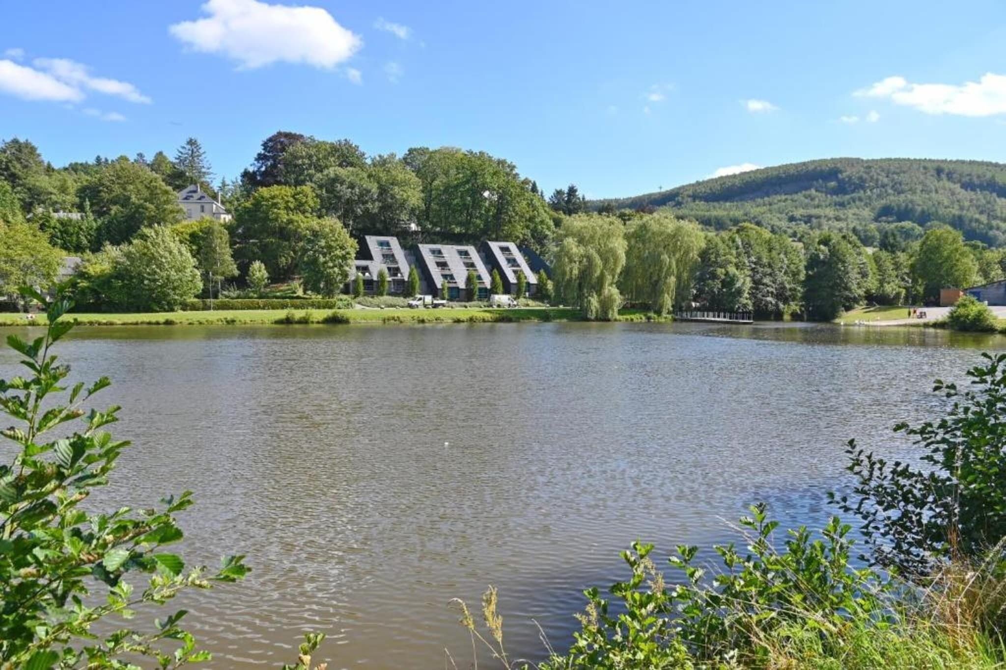 A lakeside view with a modern building, lush greenery, and a hill in the background.