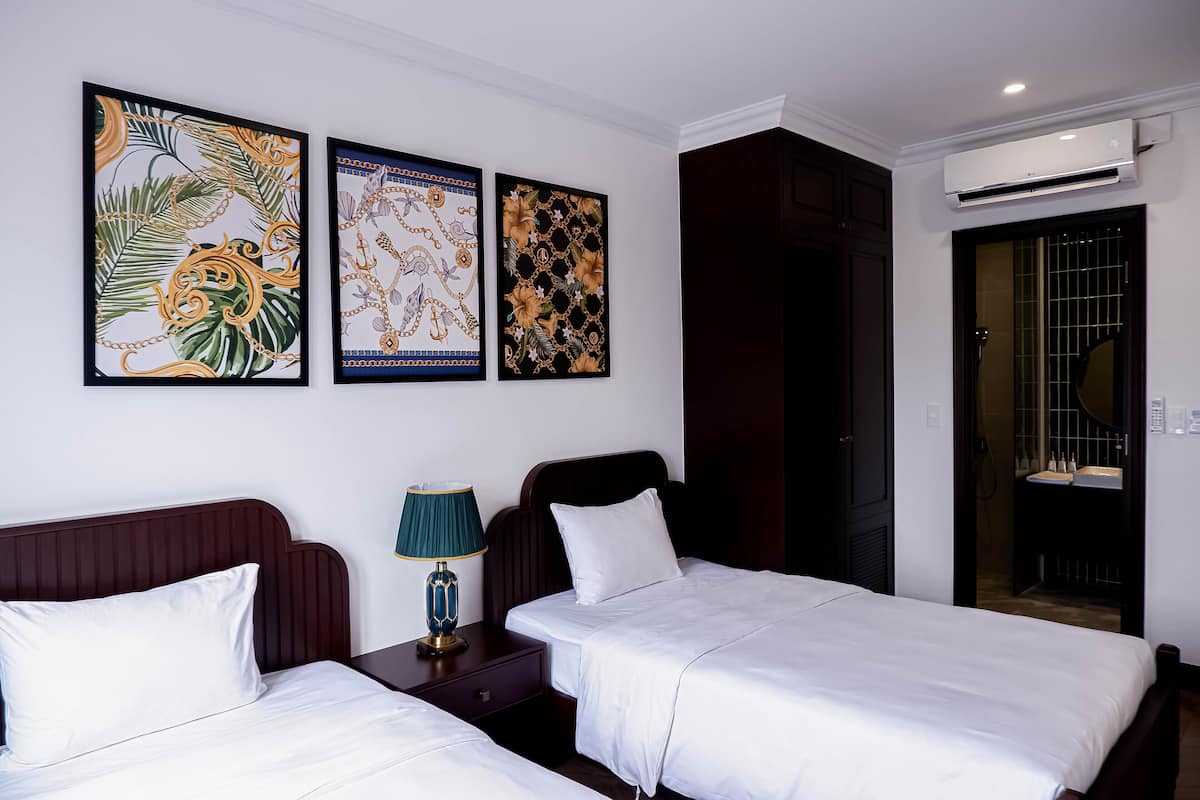 A hotel room with two beds, a nightstand, a lamp, and three framed artworks on the wall.