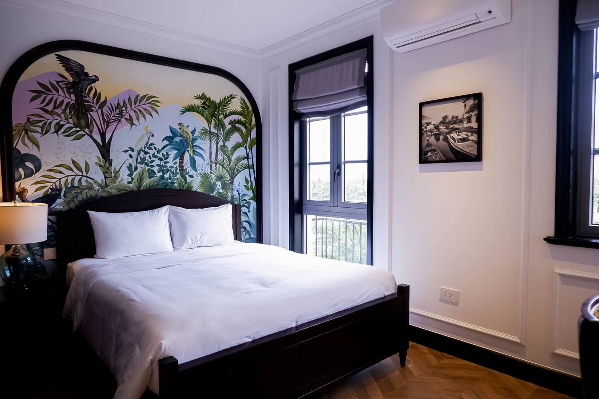 A neatly made bed with white linens, a dark headboard, and a wall mural of tropical foliage.