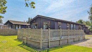A wooden cabin with a fenced deck, surrounded by grass and trees.