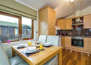 A compact kitchen with wooden cabinets, a dining table with breakfast items, and a view of a house through a window. - 2 bedroom accommodation in Carnforth (Carnforth)