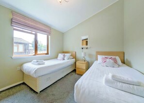 A hotel room with two beds, a window with a view of a building, a bedside table with a lamp, and a wall-mounted mirror.