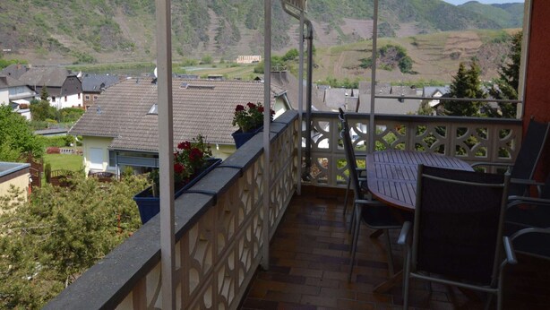 Een overdekt terras buiten met een tafel en stoelen, met uitzicht op een vallei met huizen en begroeiing.