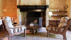 Une pièce rustique avec un mur en pierre, une cheminée, deux fauteuils rembourrés, une petite table ronde et une bibliothèque remplie de livres.