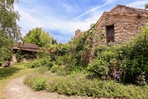 Un edificio de piedra con una ventana, rodeado de abundante vegetación y plantas florecientes.