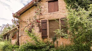 Un bâtiment rustique en pierre, doté de volets en bois, entouré d’une végétation luxuriante et de plantes en fleurs.