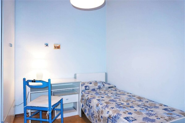 Une petite chambre avec un lit simple, un bureau et une chaise, un lit recouvert d’une courtepointe à motifs et deux petits éléments de décoration murale.