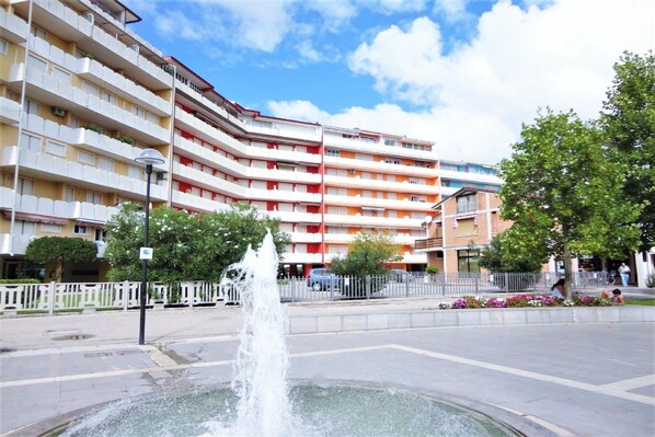 A multi-story building with balconies, a fountain, and a pedestrian area with trees and a fence. - Holiday Home in Caorle near Beach (Caorle (VE))