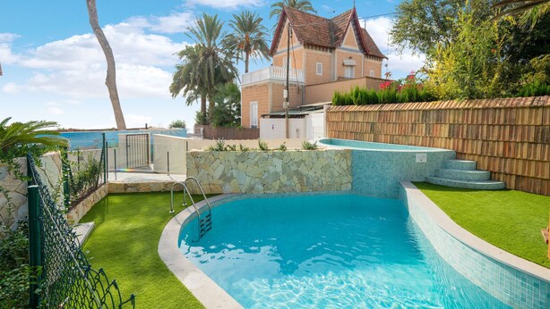 A swimming pool with a stone wall and a wooden fence, surrounded by greenery and palm trees.