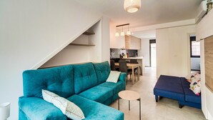 A modern living room with a teal sofa, a small round table, and a blue sofa bed. There is a staircase leading to another level, a kitchen area in the background, and a hanging light fixture.