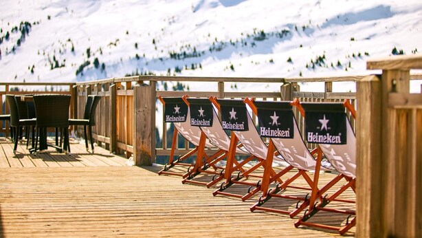 Terraza de madera con asientos y carteles de Heineken, con vista a un paisaje montañoso nevado.