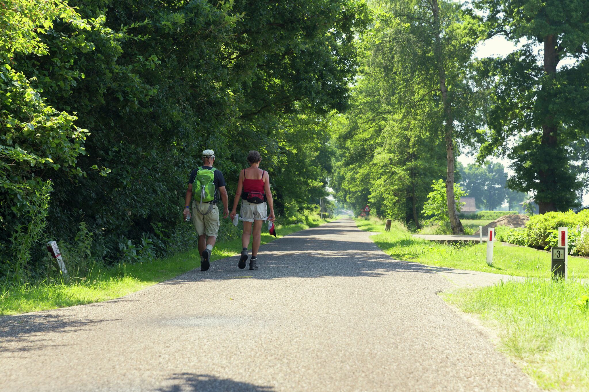 Een verharde weg langs weelderige groene beplanting, met twee personen die van de kijker weg wandelen.