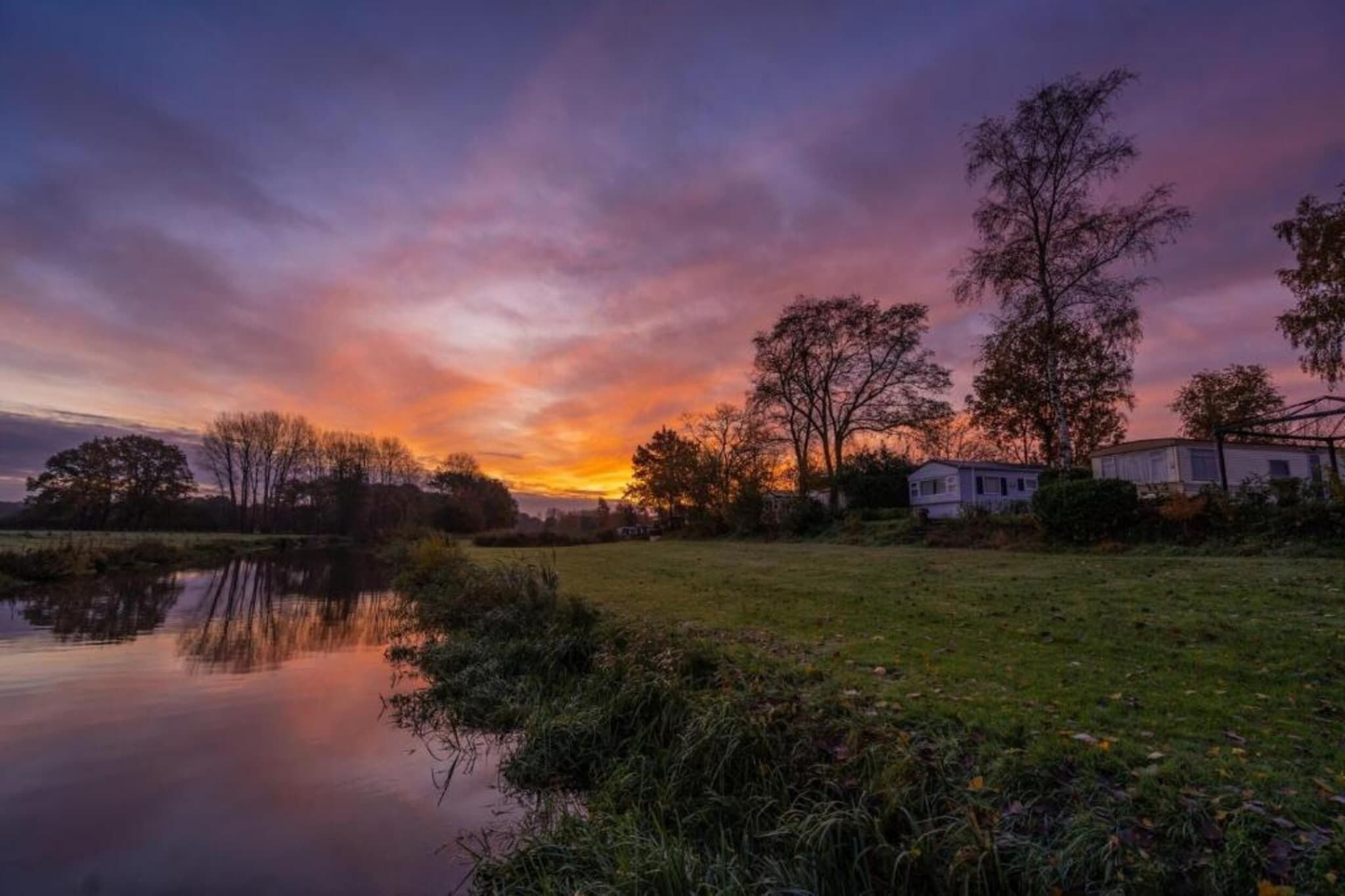 A serene lakeside scene with a sunset, trees, and a house in the background.