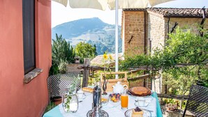 An outdoor dining area with a table set for breakfast, including bread, fruit, and a cake, under a large umbrella. - Relaxing Retreat in Le Marche (Monte San Martino)